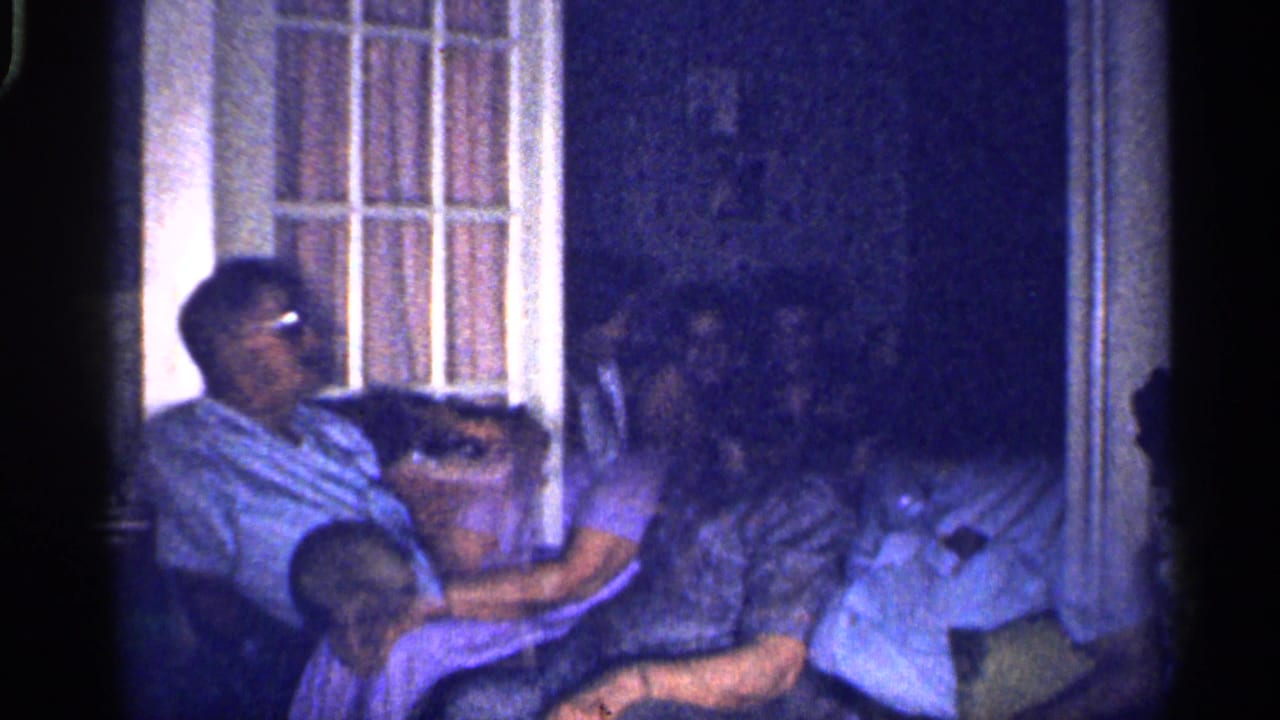 1959 Florida home-movie frame of adults seated close together in a dim interior or porch area beside a barred window.