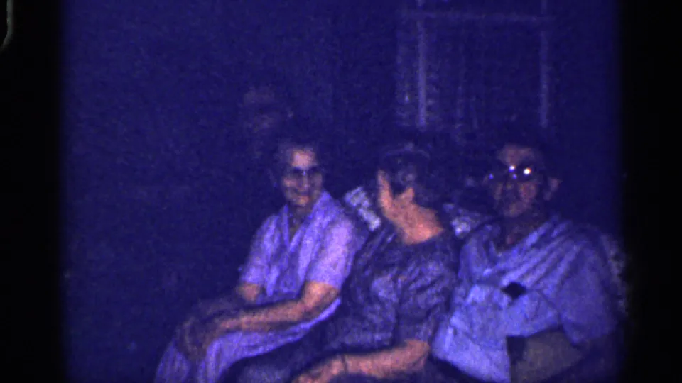 Dim 1959 Florida interior frame showing three adults seated together, partly obscured by shadow and blue film cast.