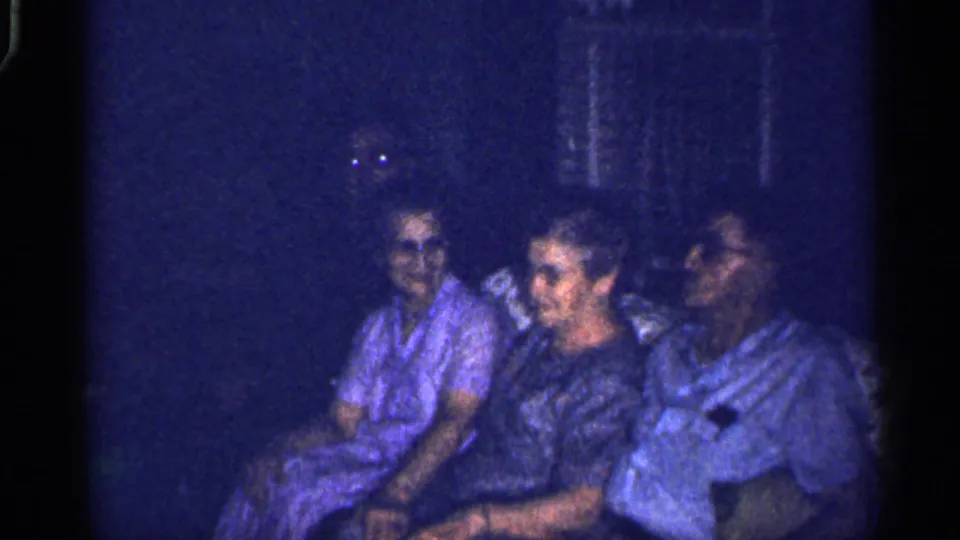 Blue-tinted 8mm frame from 1959 Florida showing seated adults in conversation or rest inside a dark domestic space.