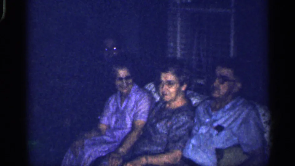 1959 Florida home-movie still of adults seated side by side in a shadowed room or porch, with a window visible behind them.