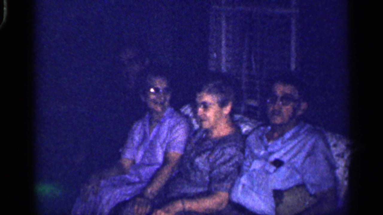 Four people seated on a couch in a dim living room, with one person turned toward the others.