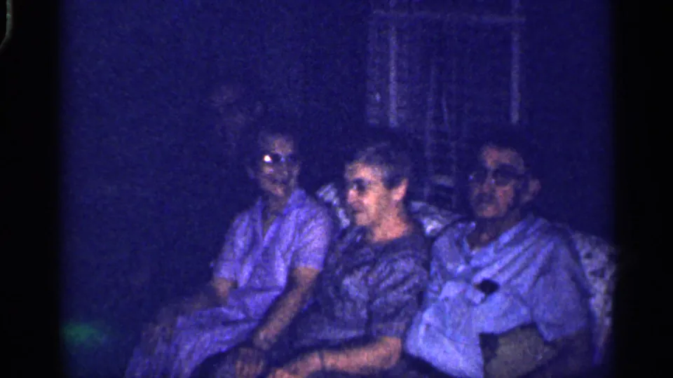 1959 Florida 8mm frame of four people seated close together on a couch in a dark living room, reacting casually to the camera.