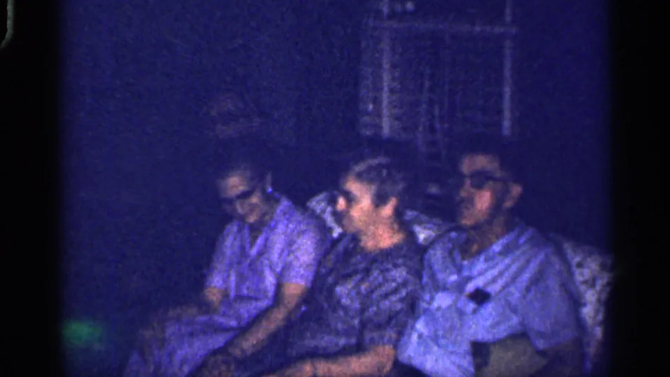 A candid 1959 Florida home-movie frame shows a small group on a couch, with faces visible in low blue-tinted light.