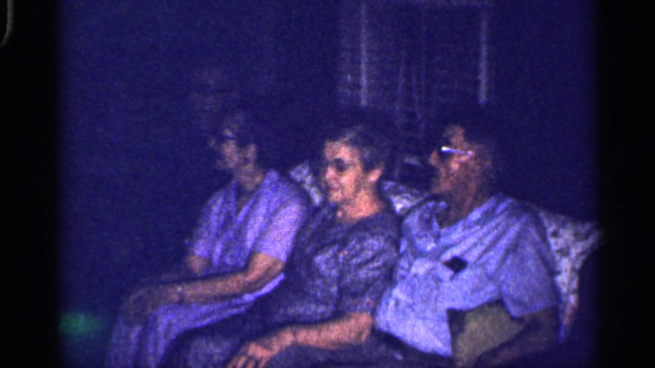 Three adults sit close together on a pale couch in a dim room.