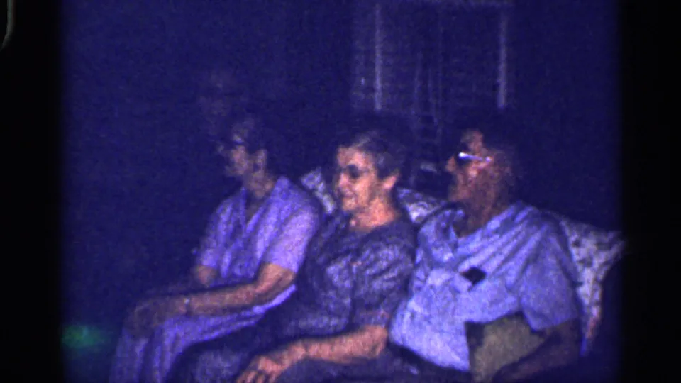 Three adults sit on a pale couch in a dim Florida home interior, with two wearing glasses and one woman turned toward the group.