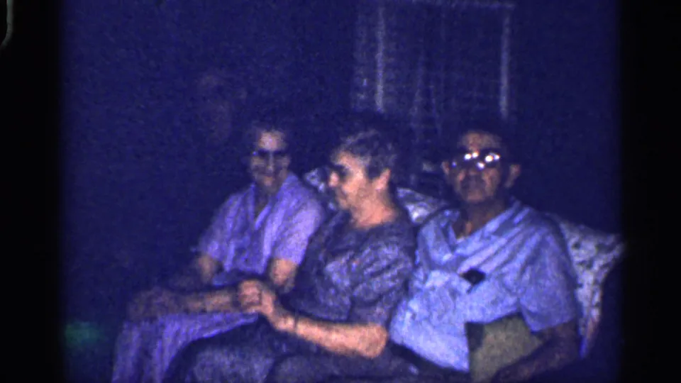 In a 1959 Florida home-movie frame, three adults sit on a couch, with the man at right wearing glasses and the center woman smiling.