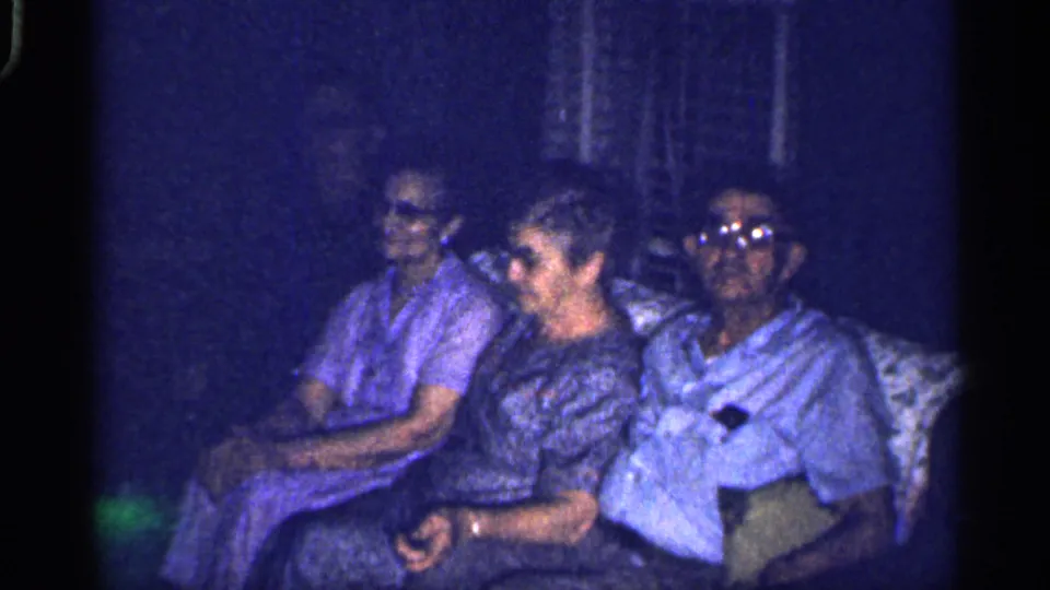 Three adults remain seated close together on a couch, with eyeglasses catching light in the dim Florida home interior.