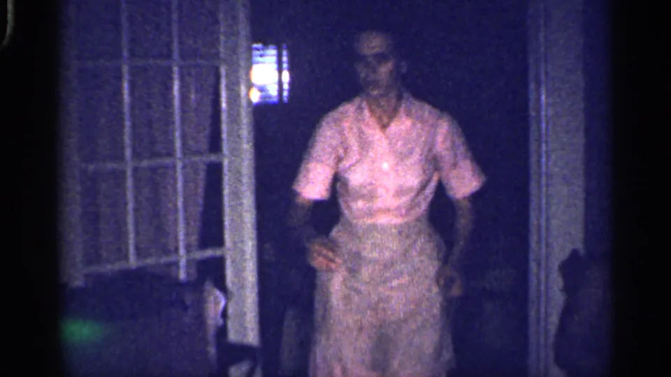 A woman in a light blouse and skirt stands near a doorway or window frame in the same dim 1959 Florida home-movie interior.