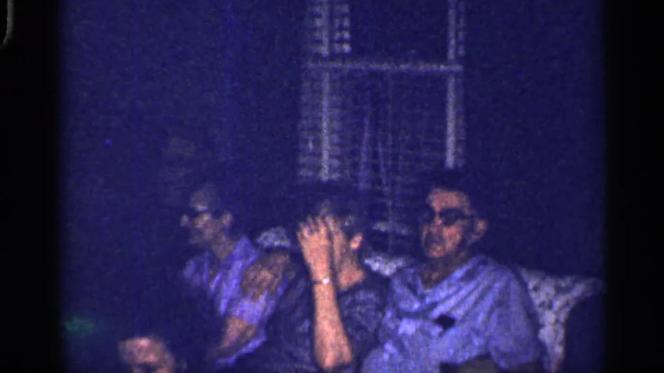 1959 Florida home-movie frame of adults seated close together on a couch in a dark living-room interior.