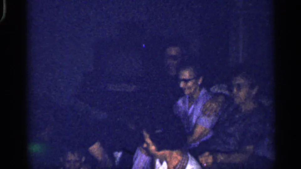 Dim 1959 Florida home-movie frame with several adults seated and turned toward one another in a private interior.