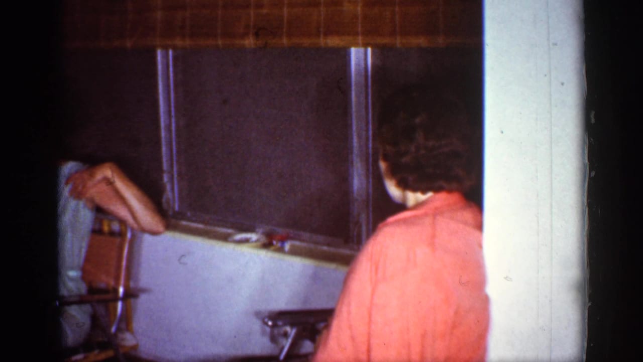 1959 Florida home-movie frame of people in a private interior, with one person seen from behind and another partly hidden by the window area.