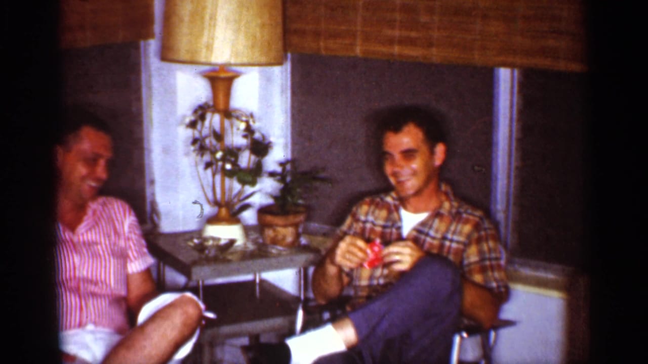 Two adult men sit in a Florida living room, laughing near a gold-shaded lamp, potted plants, and bamboo blinds.