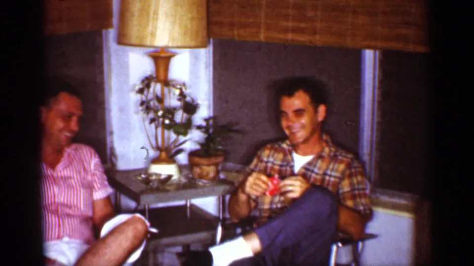 Two adult men sit in a Florida living room, laughing near a gold-shaded lamp, potted plants, and bamboo blinds.