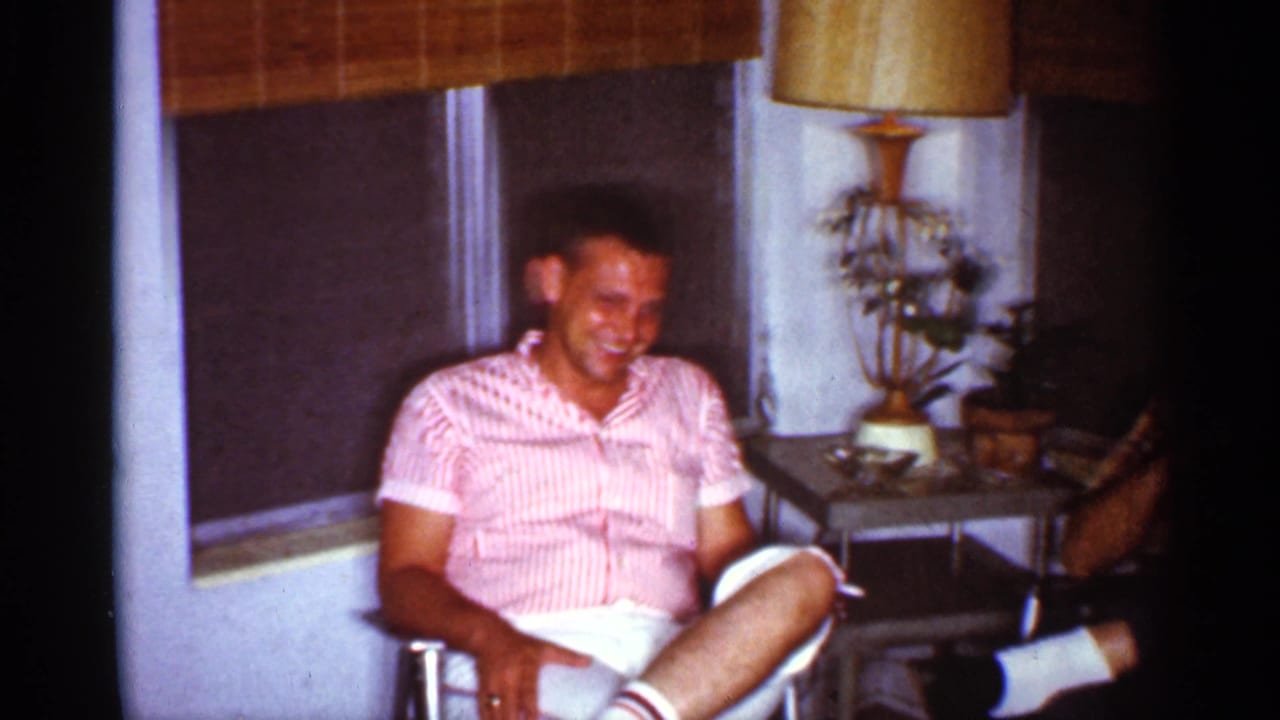 A seated man in a pink striped shirt laughs in a 1959 Florida living room beside a lamp, plants, and shaded windows.
