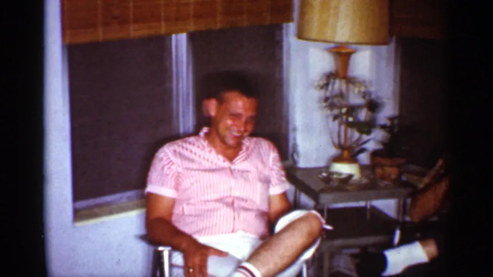 A seated man in a pink striped shirt laughs in a 1959 Florida living room beside a lamp, plants, and shaded windows.