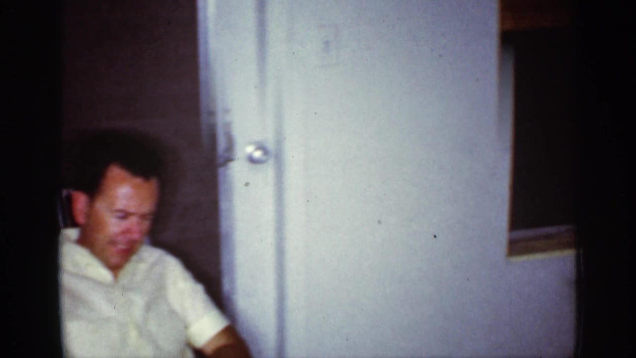 An adult man in a light shirt sits near a white door within the same dim Florida home-movie living room interior.