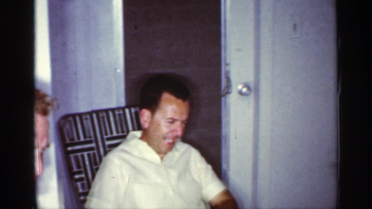 In 1959 Florida home-movie footage, two adult men in light shirts sit indoors near a doorway and patterned chair.