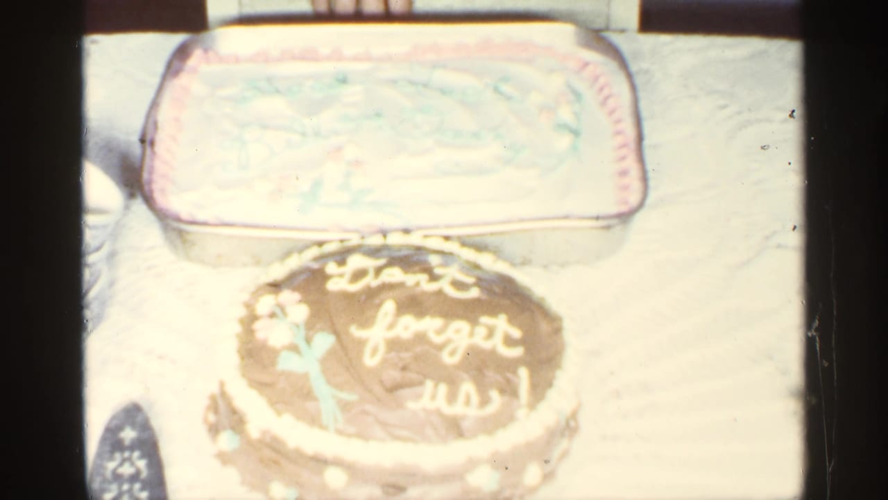 The camera frames a chocolate cake with pale icing lettering in front of a rectangular frosted cake on a table.