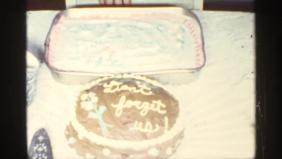 The camera frames a chocolate cake with pale icing lettering in front of a rectangular frosted cake on a table.