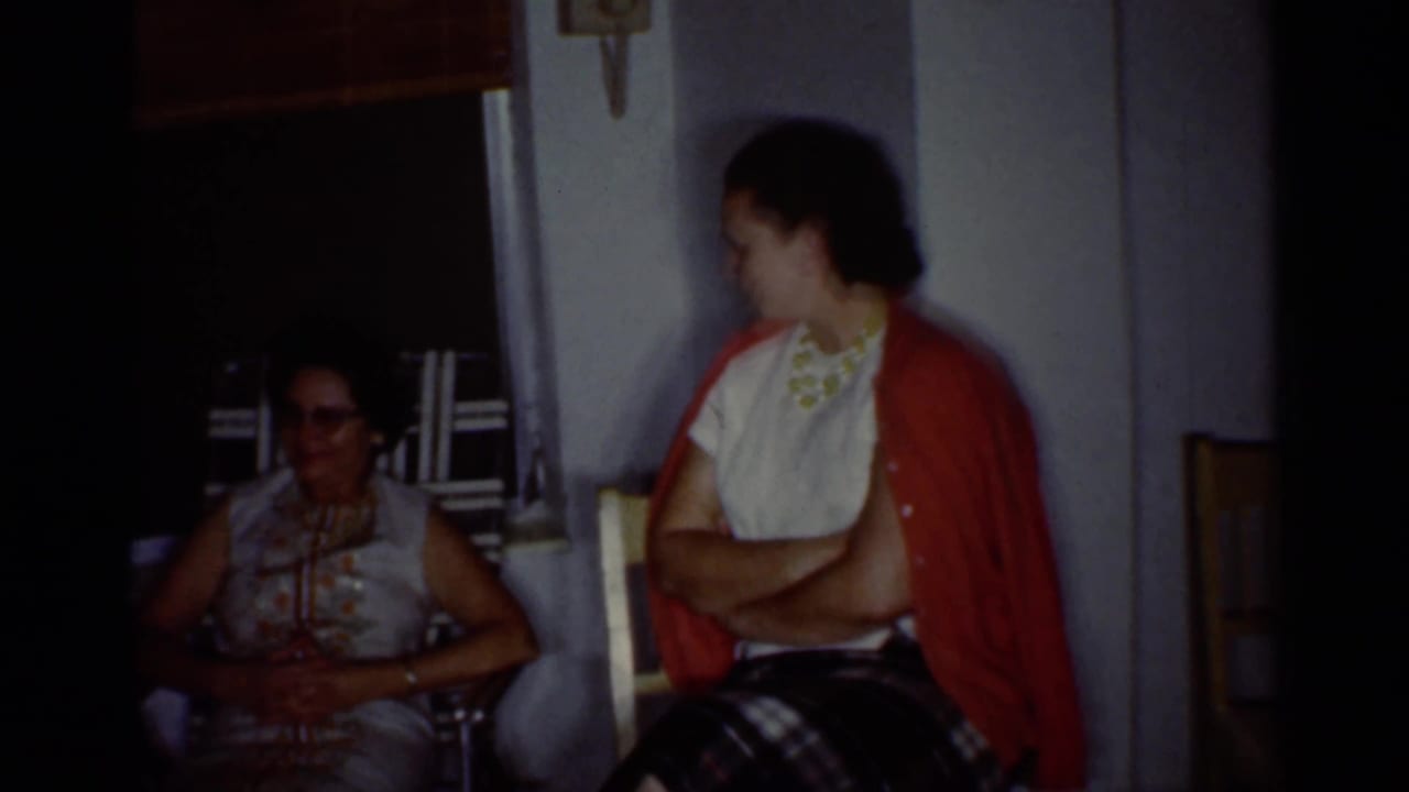 Two women seated indoors near a shaded window, one partly in shadow and one wearing a red cardigan.