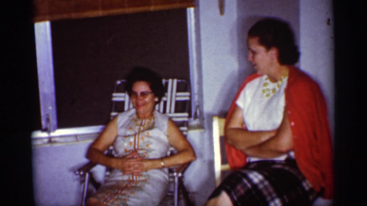 1959 Florida home-movie frame showing two women seated in a sunlit interior, with window shades behind them and casual mid-century clothing.