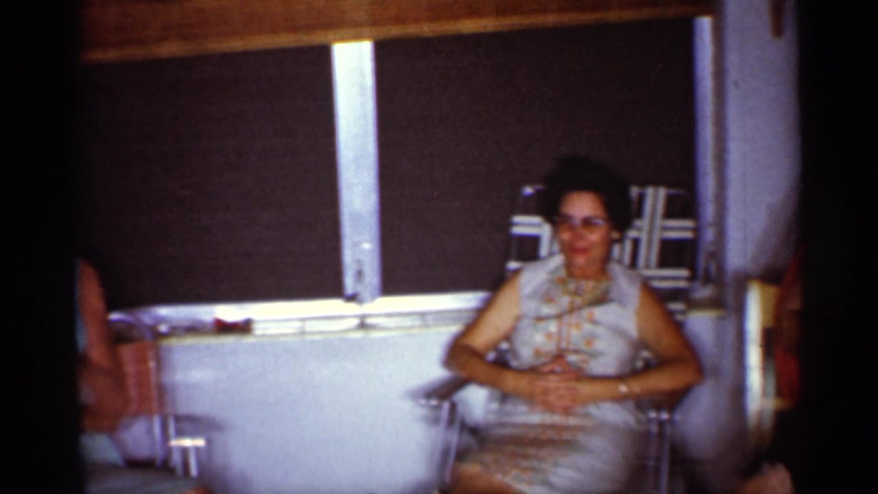 1959 Florida home-movie frame of a seated woman in sunglasses and a light patterned dress, filmed below a dark window shade indoors.