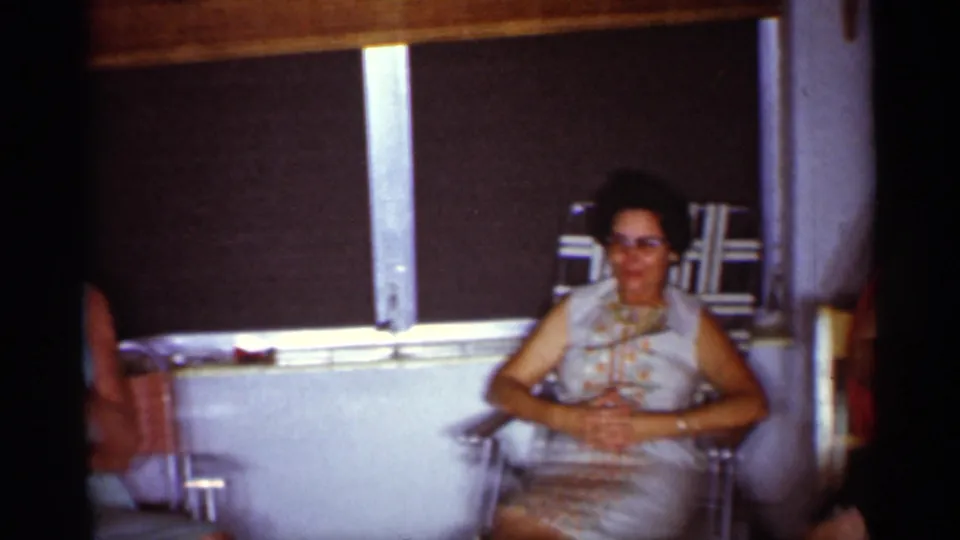1959 Florida home-movie frame of a seated woman in sunglasses and a light patterned dress, filmed below a dark window shade indoors.