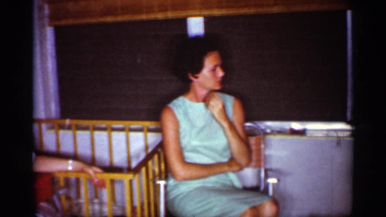 1959 Florida home-movie frame of a woman in a mint sleeveless dress seated indoors near a shaded window and wooden railing.
