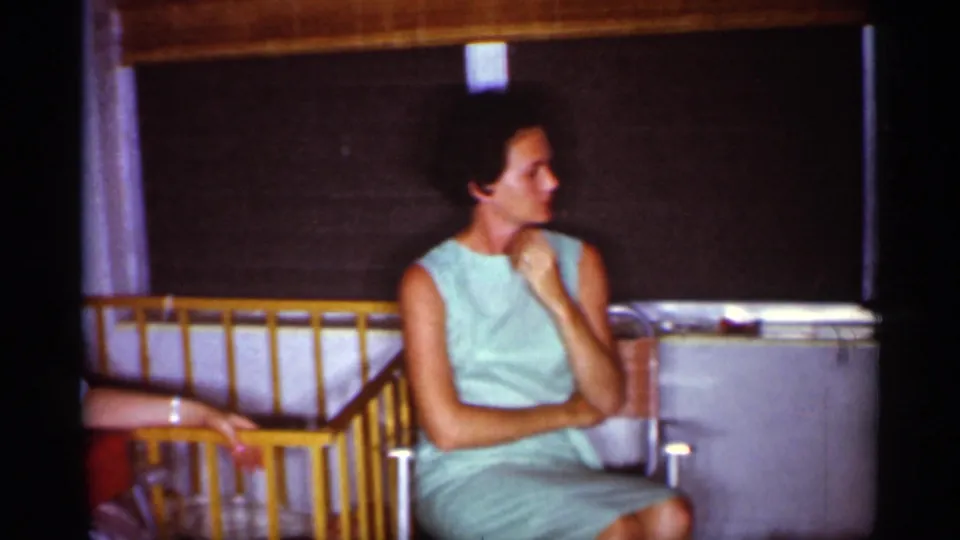 1959 Florida home-movie frame of a woman in a mint sleeveless dress seated indoors near a shaded window and wooden railing.
