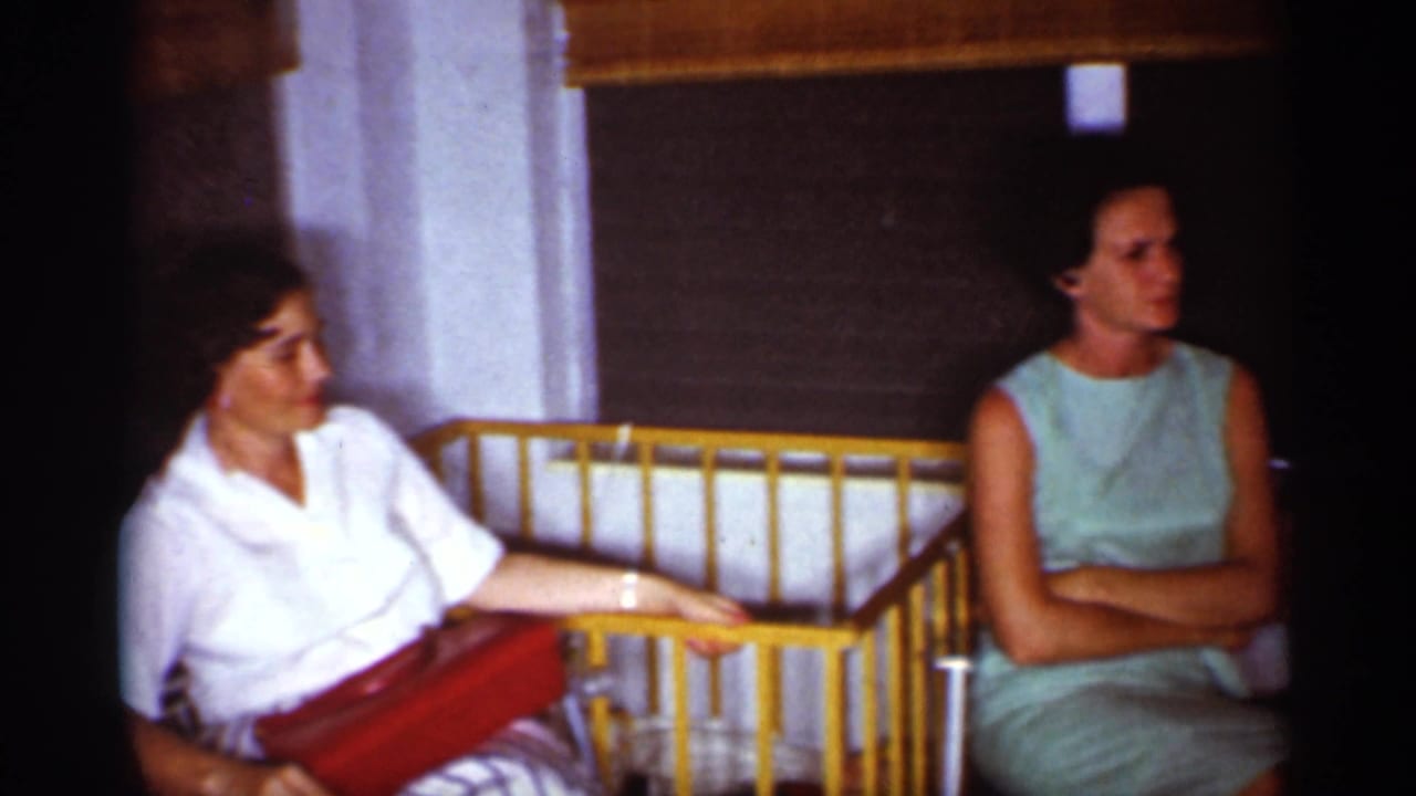 Two women seated indoors beside a yellow baby crib, one holding a red purse.