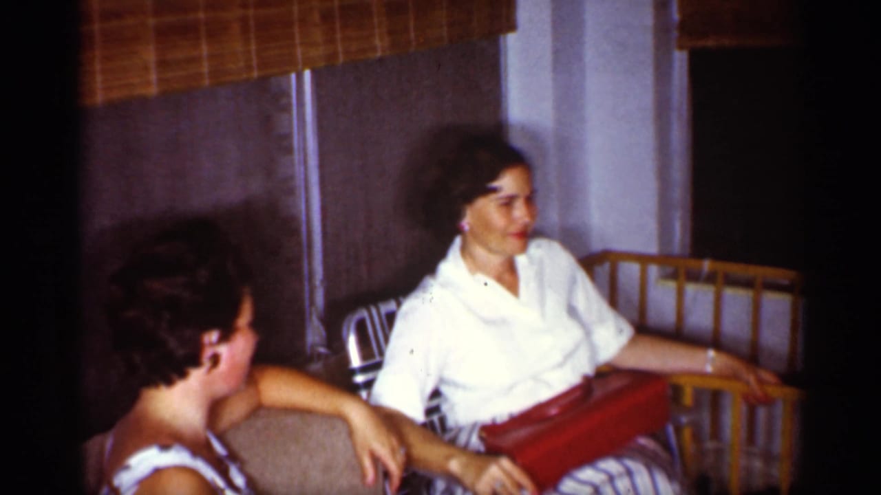 1959 Florida home-movie frame with two women seated by a baby crib, one seen partly from behind and one facing the camera side.