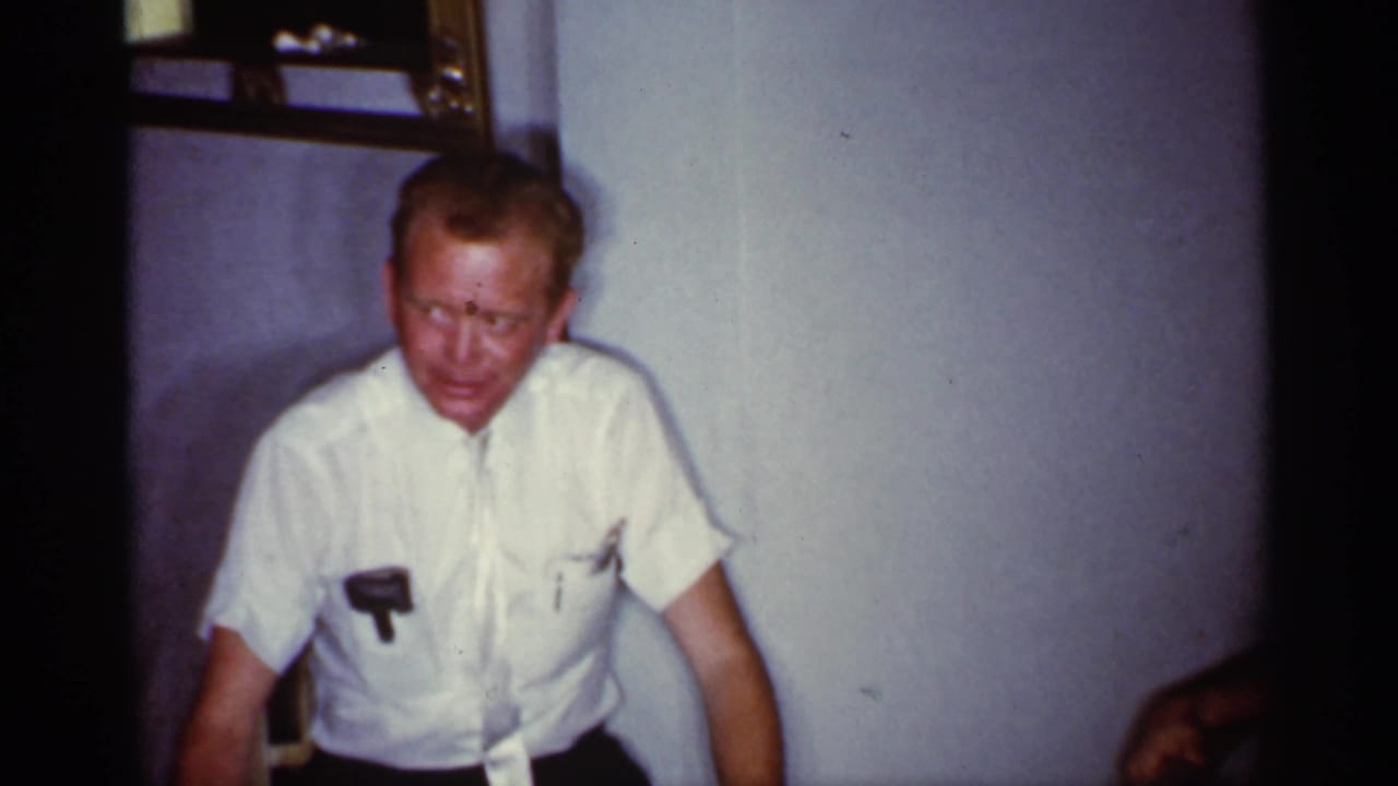 1959 Florida home-movie frame showing a seated man in a white short-sleeve shirt, softly focused against a plain wall.