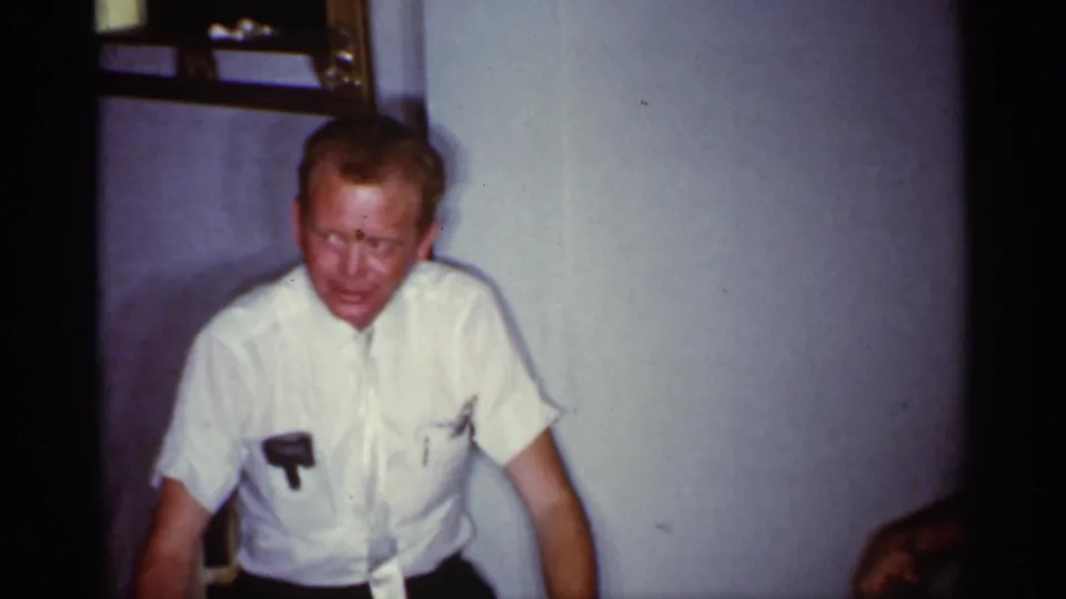 1959 Florida home-movie frame showing a seated man in a white short-sleeve shirt, softly focused against a plain wall.
