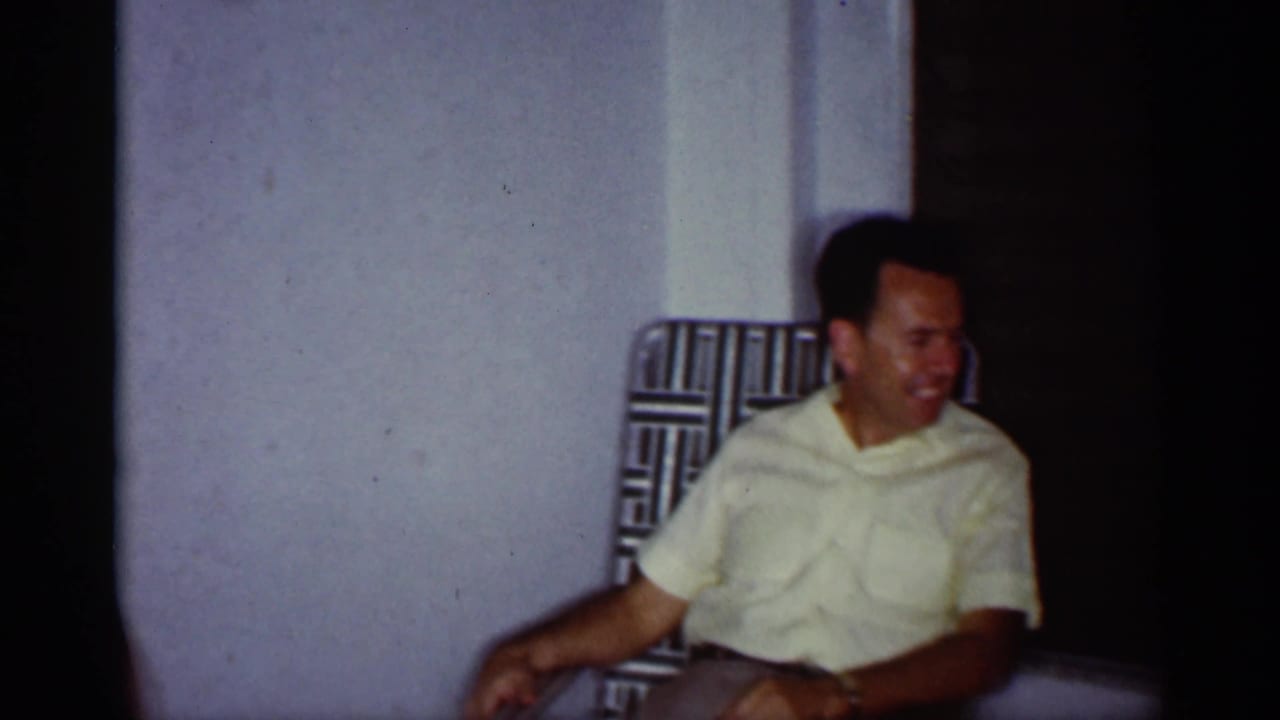 1959 Florida home-movie frame of a man seated in a patterned chair inside a modest private room, with soft 8mm color.