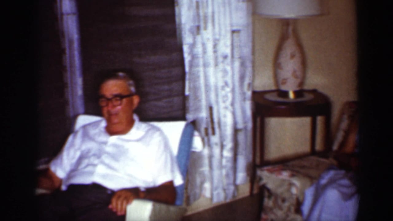 In a 1959 Florida home interior, an older man in glasses sits in a chair near lace curtains, a shaded window, and a patterned lamp.