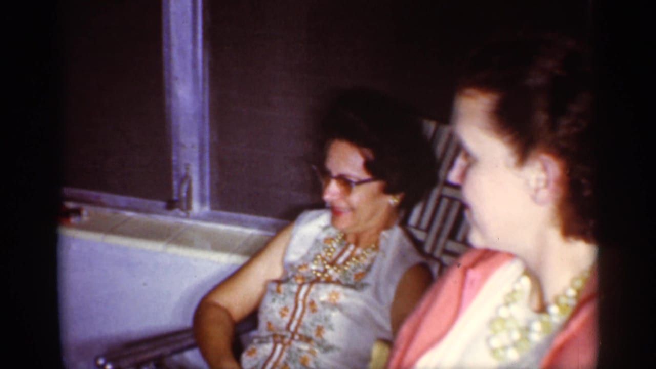 Two women seated by a window, one wearing glasses and one wearing a pearl necklace.
