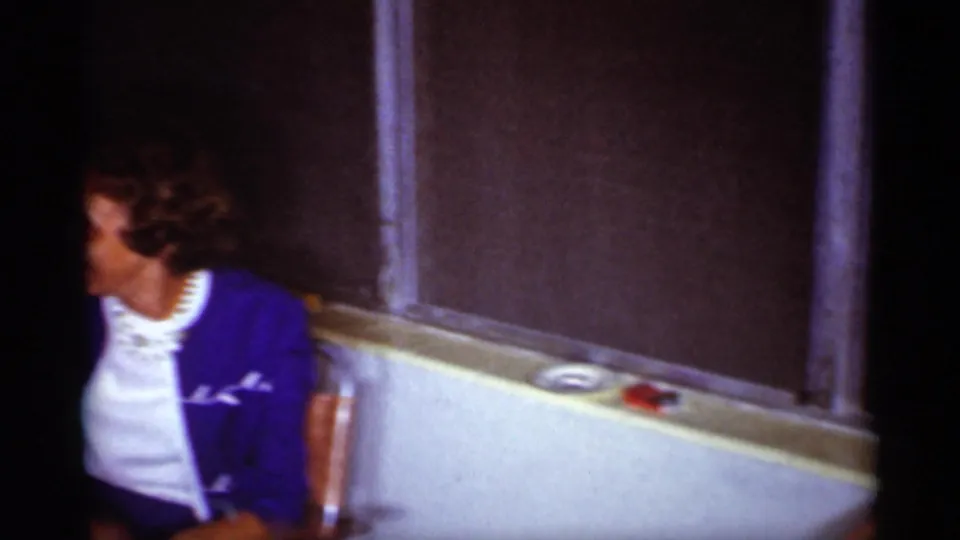 A woman in a blue jacket sits near the living room window ledge, with shades, pale wall trim, and small objects visible nearby.