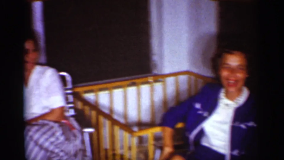 A smiling woman sits beside a wooden crib rail in the 1959 Florida living room, while another adult is partly visible in the background.