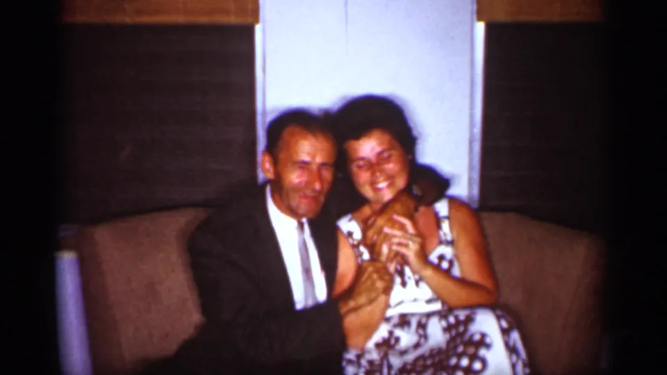The couple sits closely on a brown couch, smiling toward the camera during a 1959 Florida home-movie family gathering.