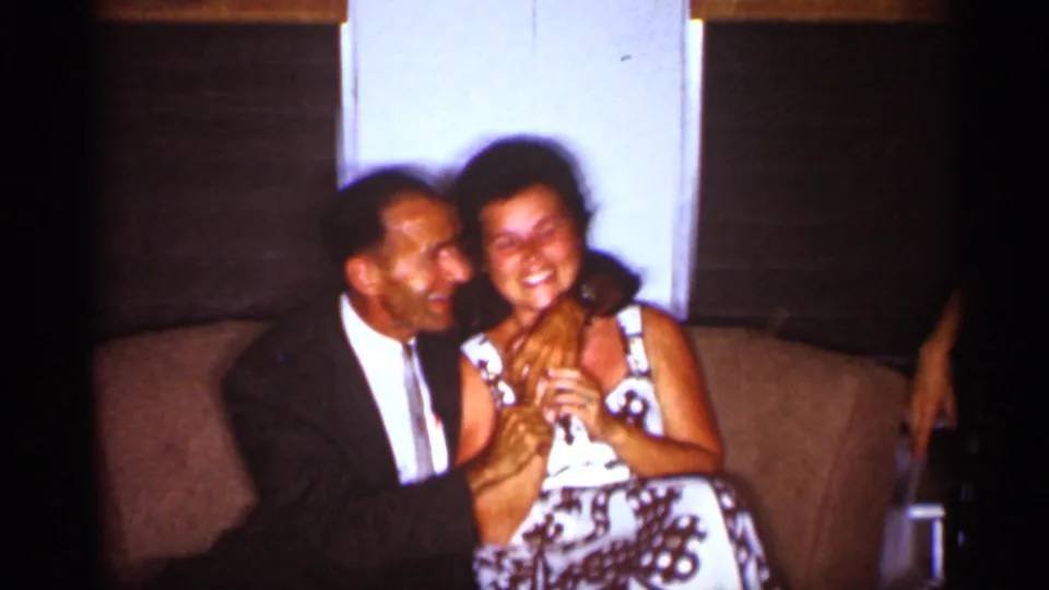 A man appears to kiss or nuzzle a smiling woman on a couch in a warm 1959 Florida home-movie living room scene.