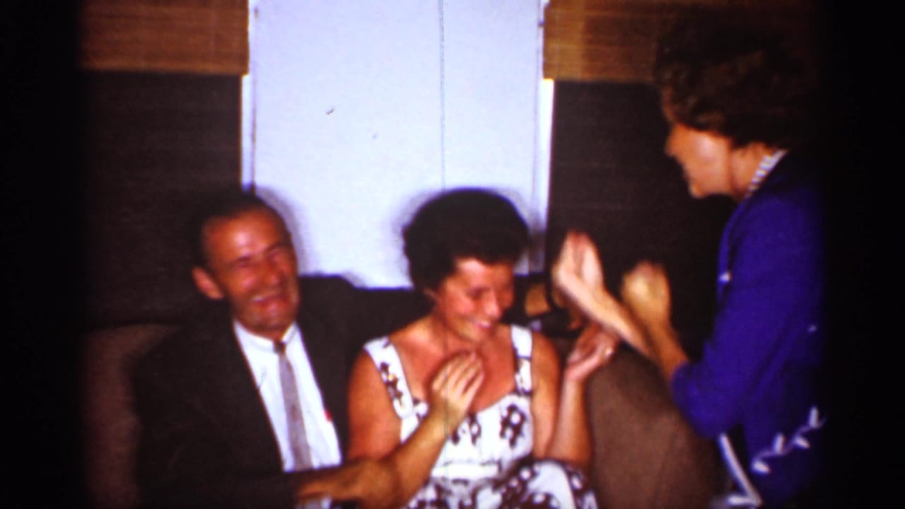 1959 Florida home-movie frame of adults in a living room, with a woman in blue engaging a seated laughing woman.