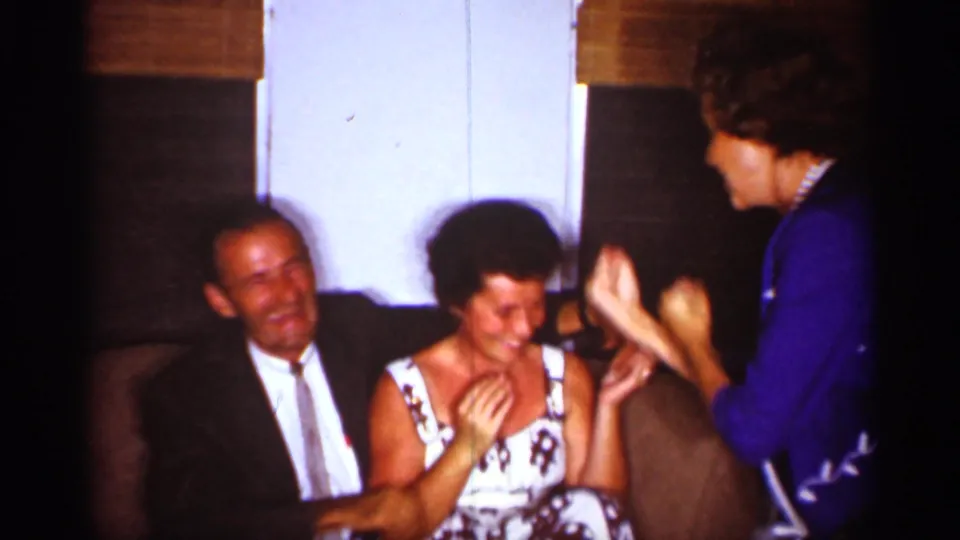 1959 Florida home-movie frame of adults in a living room, with a woman in blue engaging a seated laughing woman.