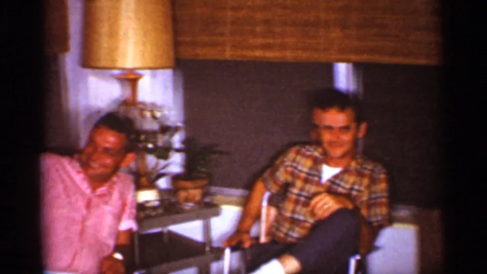 Two men relax in chairs near a table lamp and bamboo blinds in this 1959 Florida home-movie frame.
