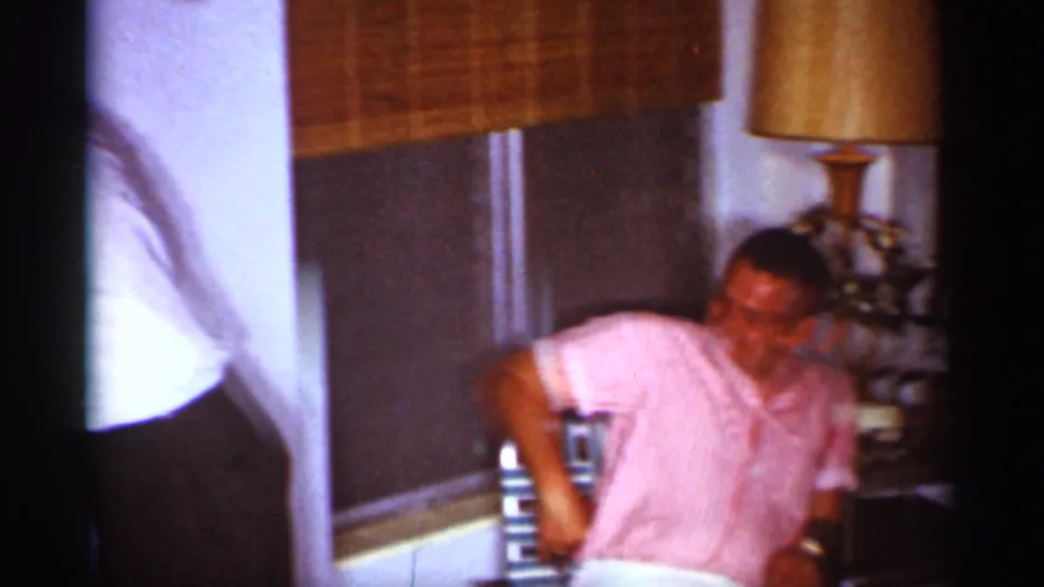 A man in a pink striped shirt sits by a table lamp and bamboo blinds in the Florida living room gathering.