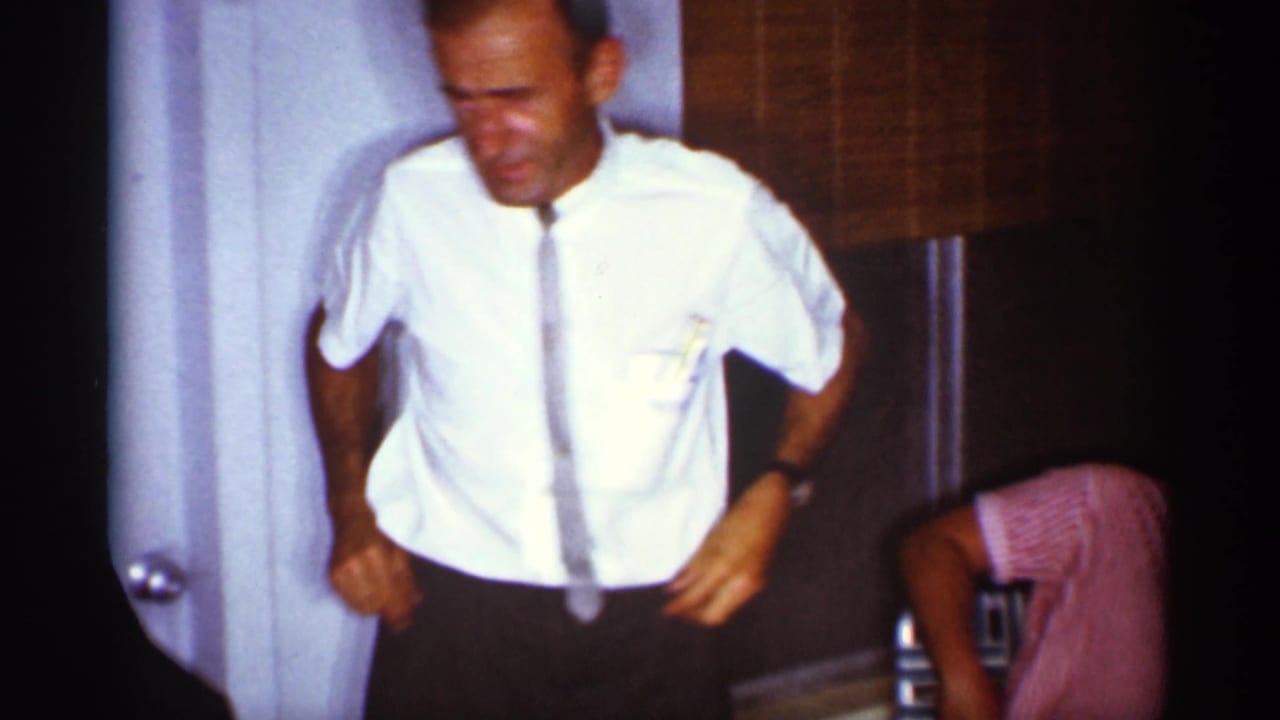 In 1959 Florida home-movie footage, a man in a white short-sleeved shirt and tie stands or leans near an interior doorway and bamboo blind.