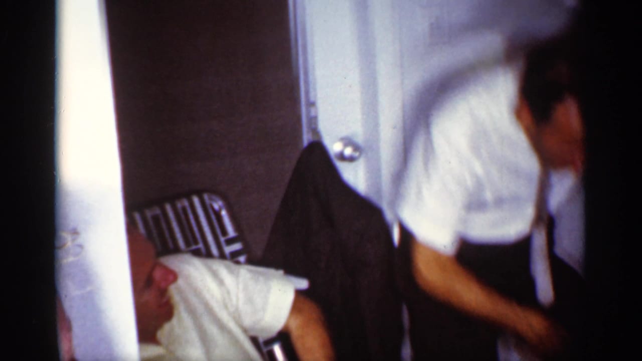 Seated man in a white shirt near a metal lawn chair while another man moves at the right edge.