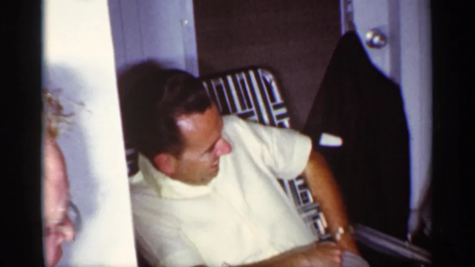 In the Florida interior, a man in a white shirt sits in a metal lawn chair beside a doorway, with a dark coat hanging nearby.