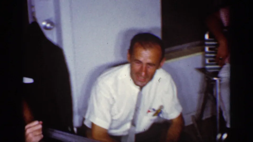 A man in a white short-sleeved shirt and tie squats or leans forward in the 1959 Florida interior, near a door and hanging coat.