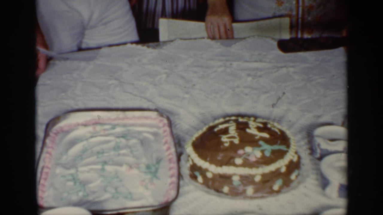 Two decorated cakes on a table with adult hands and torsos nearby.
