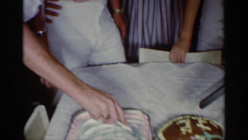 A close home-movie view shows an adult hand beginning to cut the sheet cake next to the round birthday cake.
